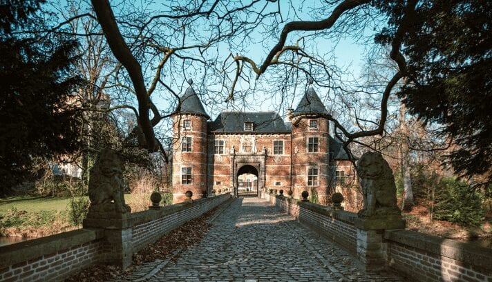A large castle in Belgium with tall towers and stone walls, surrounded by lots of green trees and plants.