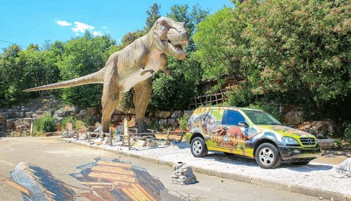 Dinosaur statues at Dinopark Funtana, surrounded by plants, and the park's car make it look like a scene from a long time ago.