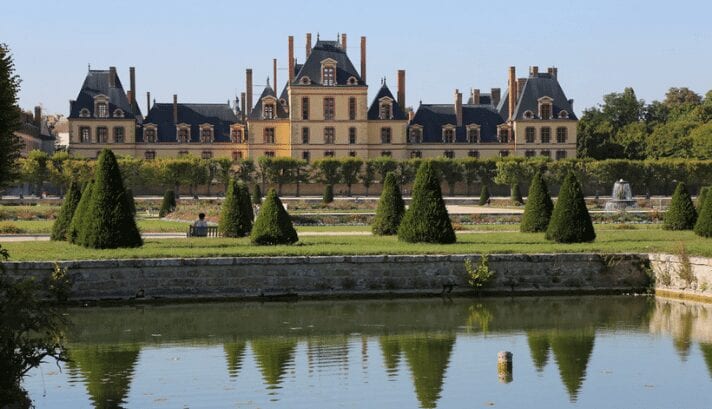 The Kings and Queens Promenade at Chateau de Fontainebleau, with elegant statues and gardens alongside a peaceful lake, reflecting the grandeur of the French palace.