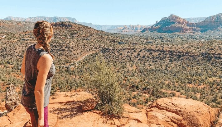 "A woman standing at a high viewpoint, gazing at the red rock formations and greenery of Sedona below a clear, blue sky.