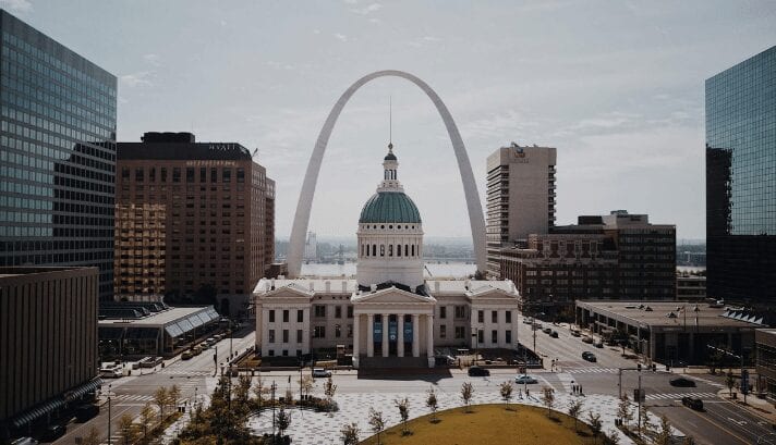 A photo of the St. Louis skyline, including the iconic Gateway Arch, with buildings and parks under a bright sky.