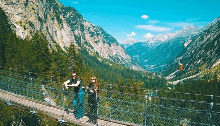 A man and woman were posing together in the middle of a hanging bridge in Switzerland, with the scenic mountains as a picturesque backdrop.