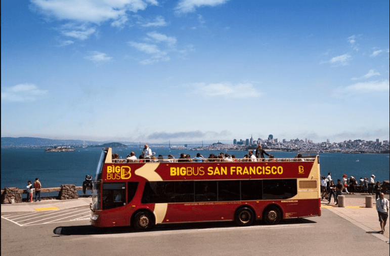 A red bus filled with tourists, traveling along a scenic route, with passengers enjoying the sights of San Francisco.