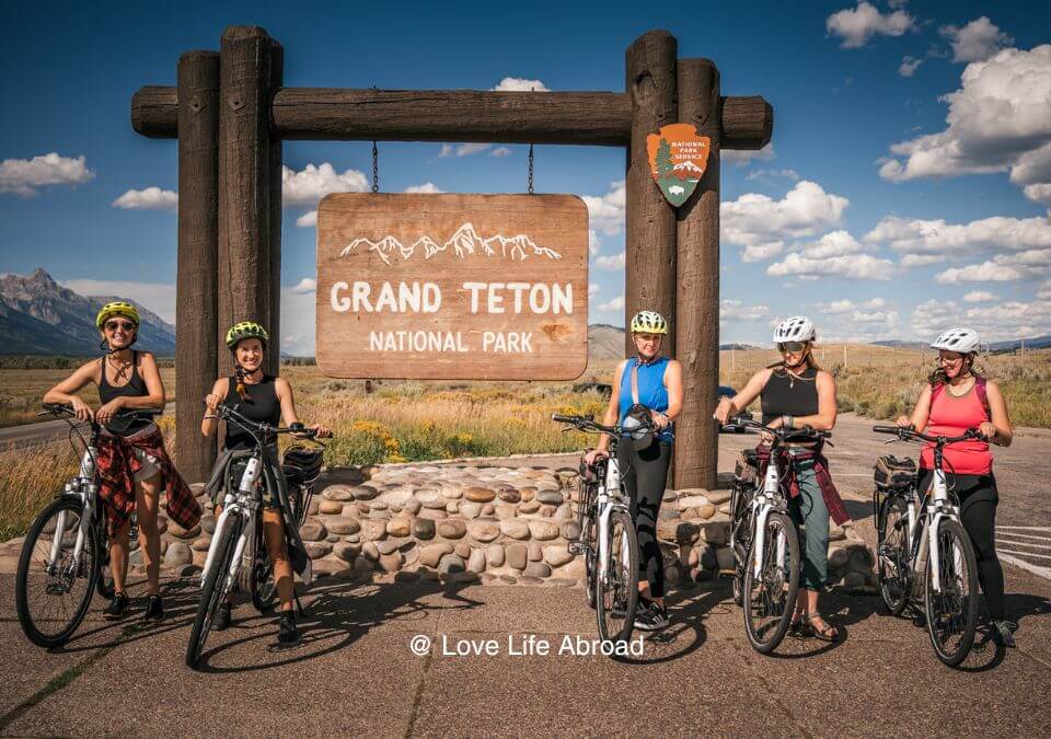 Friends biking to the Grand Teton National Park sign Photo credit wechooseadventures